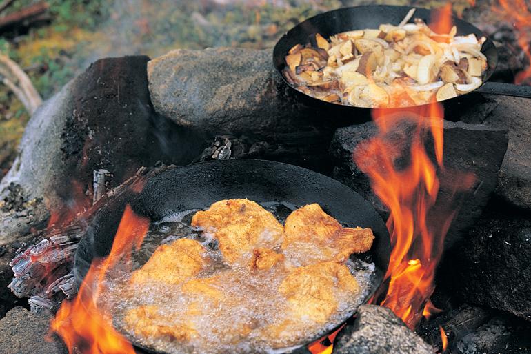 Two cast iron pans cooking over an open campfire with flames rising around the rocks